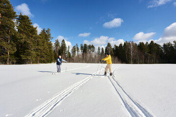 Couple in 50s pauses during cross country skiing, husband leans on poles, woman does too, both...