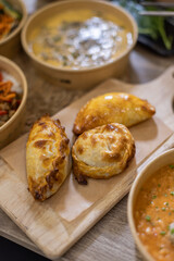 Vertical close-up of three golden-baked empanadas served on a wooden board lined with parchment paper. Warm tones, shallow depth of field, rustic food setting. Copy-space