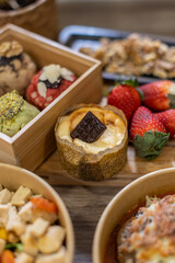 Vertical close-up of a mini cheesecake topped with a square of dark chocolate. Dessert is set among fresh strawberries and assorted cookies in a cozy, rustic food styling setup. Gourmet takeaway meal.