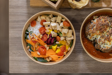 Top-down view of a vibrant Colorful vegan rice bowl served in kraft container with white rice, tofu cubes, cherry tomatoes, shredded carrot. Balanced and vibrant food-to-go, in plastic-free packaging.