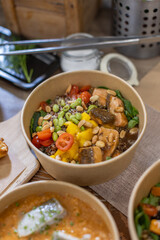 A vibrant poke bowl featuring glazed salmon, mango chunks, cherry tomatoes, edamame, peanuts, and quinoa over spinach. Vertical close-up on wooden table. Takeaway, environmentally friendly packaging