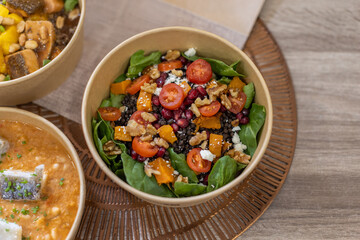 A healthy salad with black lentils, spinach, cherry tomatoes, walnuts, pomegranate, pumpkin, and feta. Horizontal close-up on a wooden surface. Healthy grab-and-go concept, biodegradable packaging