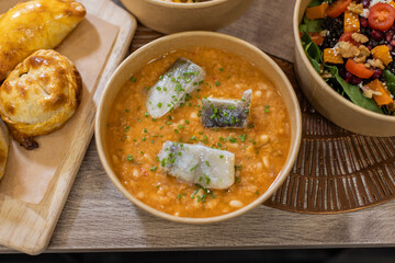A rustic Spanish-style stew with salted cod and white beans in tomato sauce, garnished with chives. Horizontal close-up shot on a wooden surface, takeaway restaurant concept, eco-friendly packaging
