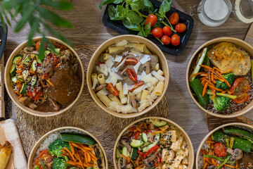  Top-down view of assorted takeaway food bowls arranged on a wooden table. Includes vegetables, pasta, sauces and fresh ingredients. Horizontal image, overhead shot, copy-space. © BASILICOSTUDIO STOCK