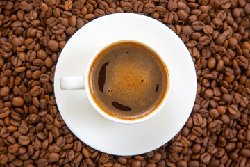 Top view of a steaming espresso in a white cup, its golden crema contrasting with the surrounding roasted coffee beans, all resting on a simple white saucer.