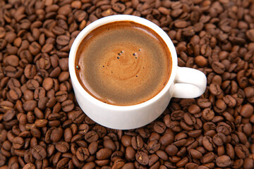 An overhead view showcasing a small white cup of espresso with a creamy foam, set on a saucer and encircled by a rustic backdrop of dark roasted coffee beans