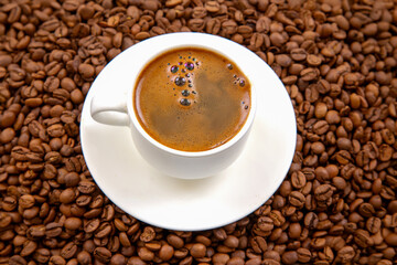 Top-down view of a white espresso cup with dark coffee and crema, placed on a matching saucer, surrounded by roasted coffee beans on a textured brown background