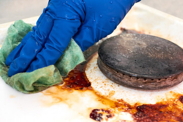 A hand in a blue rubber glove wipes a gas stove covered in grease with a rag. Cleaning a dirty...
