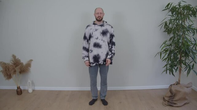 A guy in gray pants and a sweater stands against a wall, speaks and slaps himself on the head