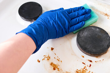 A hand in a blue rubber glove wipes a gas stove covered in grease with a scouring pad. Cleaning a dirty kitchen stove, close-up.