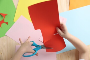 Little girl making handmade project with color paper and scissors at wooden table, top view