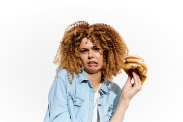 Young woman with curly hair showing disgust while holding an unhealthy burger against a white...