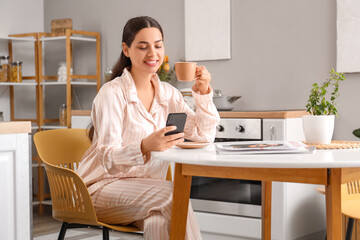 Pretty young woman with cup of hot coffee and mobile phone at home