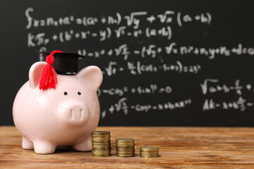 Piggy bank with graduation hat and coins on table near blackboard. Student loan concept