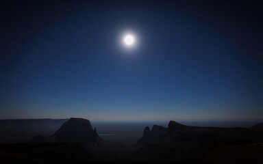 A bright sun shines over a dark, hilly landscape.  The terrain is silhouetted against a clear, blue sky, creating a dramatic contrast of light and shadow in this scenic vista.