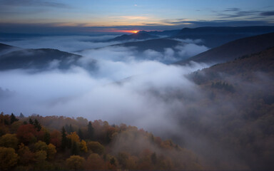 Obraz premium Misty mountain landscape at sunset. Fog blankets valleys and autumn trees on hillsides. Golden light streaks across the horizon creating a serene scenic view.