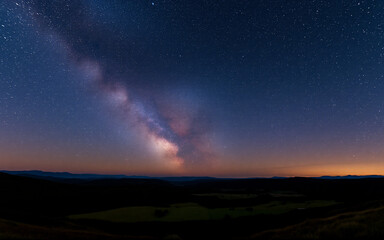 A stunning view of the Milky Way galaxy illuminating the night sky above a dark, silhouetted landscape. The cosmic scene evokes a sense of wonder and the vastness of space.