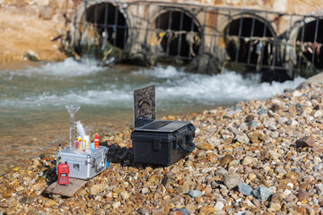 A portable water testing station with lab equipment, chemicals, and a laptop is set up beside a drainage outlet to analyze water quality and detect pollution or contamination in the environment.