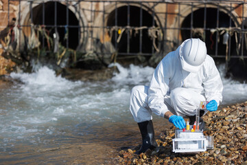 A scientist in full protective gear collects water samples from a drainage area for chemical...
