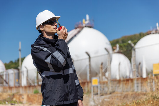 A safety officer in dark protective clothing and a white hard hat uses a walkie-talkie at a gas storage facility. He monitors the site operations near spherical tanks under a clear blue sky.