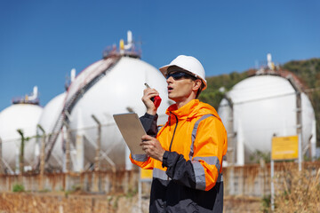 An industrial engineer in safety gear communicates via radio while inspecting spherical gas storage tanks. He holds a tablet for data logging, standing in a secured refinery or processing plant area.