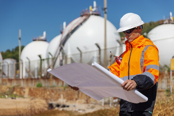 A construction engineer in protective gear examines blueprints at a gas storage facility. He holds...