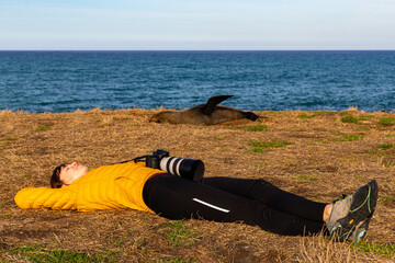 Relaxed girl lying on the grass near New Zealand fur seals by Katiki Point Lighthouse, Moeraki...