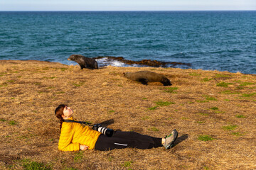Relaxed girl lying on the grass near New Zealand fur seals by Katiki Point Lighthouse, Moeraki...