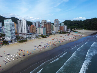 Aerial view of Praia Grande, Sao Paulo, Brazil, showing the coastline with waves reaching the sandy beach, high-rise buildings, and lush green hills in the background under a blue sky
