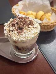 Close-up of a creamy chocolate dessert topped with whipped cream and chocolate shavings served in a glass cup on a wooden table in a café setting