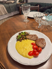 Delicious homemade dinner featuring grilled beef steak, creamy polenta, sautéed mushrooms with scallions, and cherry tomatoes served on a white plate over a wooden dining table in a cozy home setting.