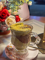 Delicious hot chocolate topped with creamy pistachio served in a transparent glass cup on a floral saucer, with decorative flowers in the background