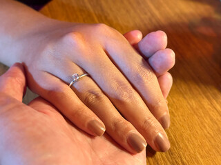 Romantic close-up of a couple holding hands, showing an elegant engagement ring with diamond on a woman’s hand over a wooden table under warm light