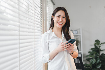 Obraz premium Cheerful woman in white shirt holding smartphone, standing by window with blinds in modern, bright interior.