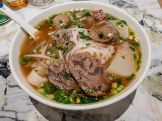 A bowl of Ho Fun Noodles Soup with beef slices, meat balls and mooli, garnished with slices onions and chopped spring onions.