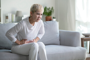Obraz premium An elderly Asian woman is sitting on the sofa at home, holding her stomach with her hands. Feels the pain of internal organs, indigestion, poisoning