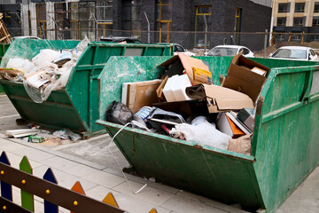 Overflowing green construction dumpster filled with cardboard boxes and building waste at urban...