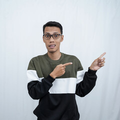 A young Asian man with glasses wearing a sweater smiles happily while pointing to the side of an empty space. Isolated on a white background.