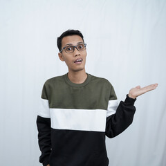 A young Asian man with glasses wearing a sweater smiles happily while pointing to the side of an empty space. Isolated on a white background.