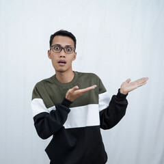 A young Asian man with glasses wearing a sweater with a shocked, surprised expression while pointing to the side of the empty space. Isolated on a white background.