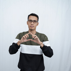 A young Asian man with glasses wearing a sweater smiles while making a love sign with his hands. Isolated on white background.