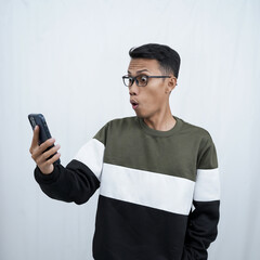 A young Asian man with glasses wearing a sweater with a surprised happy expression getting a surprise while looking at a smartphone. Isolated on a white background.