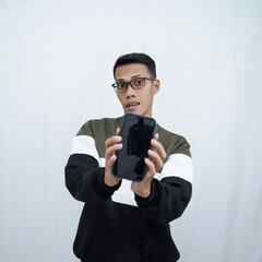 A young Asian man with glasses wearing a sweater points to a smartphone screen. Isolated on a white background.