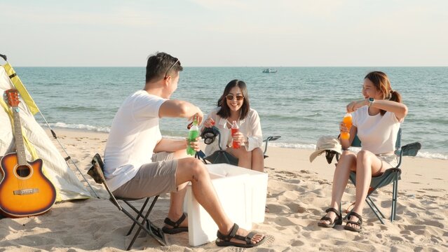 Happy friends with drinks in camp and laughing together in holidays on sand beach near camping tent vacation time at sunset, Young Asian group woman and man in summer travel outdoor
