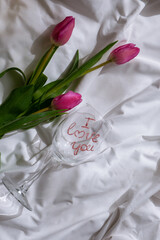 Wine glass with words I LOVE YOU Fresh bouquet of pink tulip flowers on bed. Gift present idea for holiday birthday 8 March Mother's Day Valentine's day Women's Day. Copy space