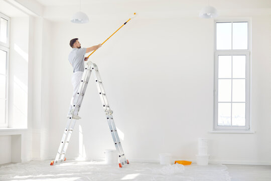 Portrait of young man painter from repair service in uniform standing on a ladder in empty room and painting the ceiling with paint roller. Renovation and renovating house concept.