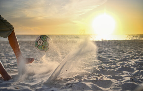 Boy kicking ball on beach towards beautiful sunset on the horizon