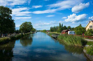 A scenic view of G&ouml;ta Canal with calm waters reflecting a bright blue sky and white clouds. Lush greenery, traditional Swedish houses, and a peaceful summer atmosphere. T&ouml;reboda, Sweden