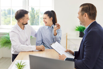 Portrait of happy smiling young married couple sitting in office with male realtor holding house...