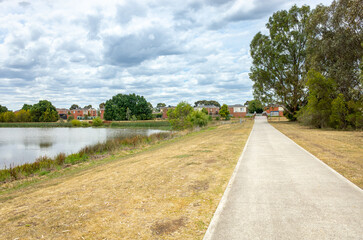 A straight paved walking path or trail in a public park running alongside Taylors Lake, Melbourne, Australia, with the concrete trail leading to suburban houses.